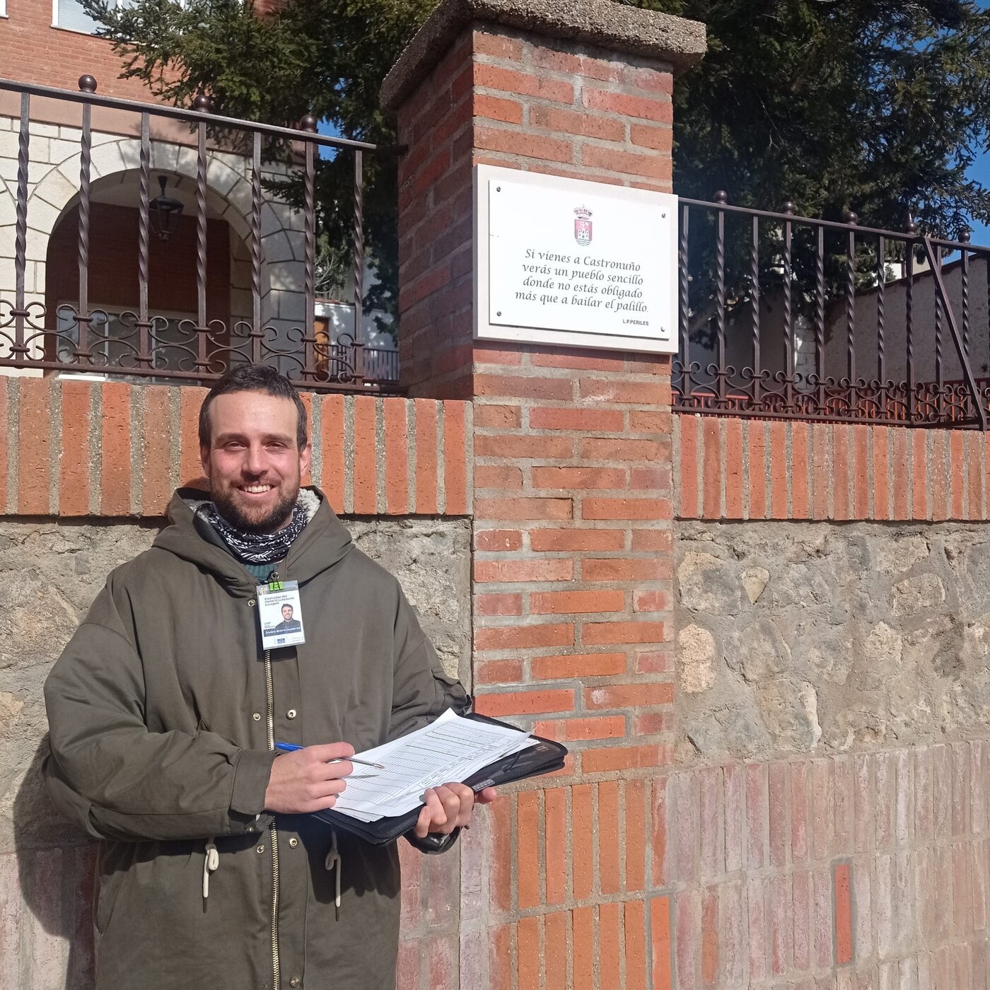 Reclutador de Marea Deliberativa con su carpeta de invitaciones, frente a la placa del municipio de Castronuño (Valladolid).
